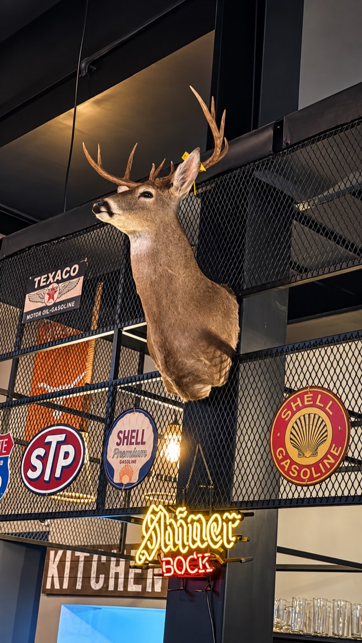 Venado disecado entre letreros vintage de gasolineras y un neón de cerveza
